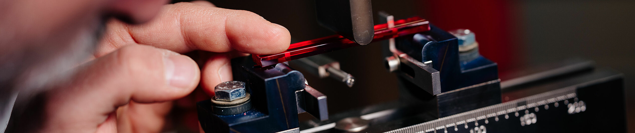 A scientist prepares to measure the deflection strength of a red colored transparent plastic strip in an apparatus