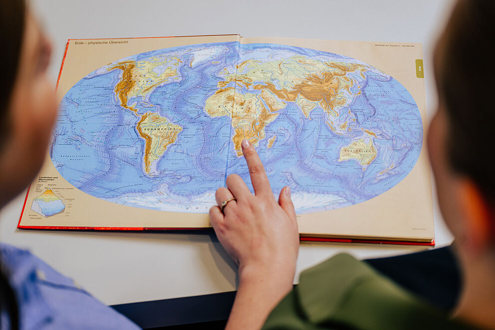 Two people are looking at an atlas with a world map, one person is pointing to the southern hemisphere with his index finger.