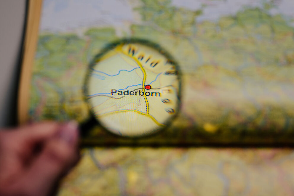 A hand holds a magnifying glass in front of an atlas, the city of Paderborn is in the centre of the magnifying glass and is highlighted.