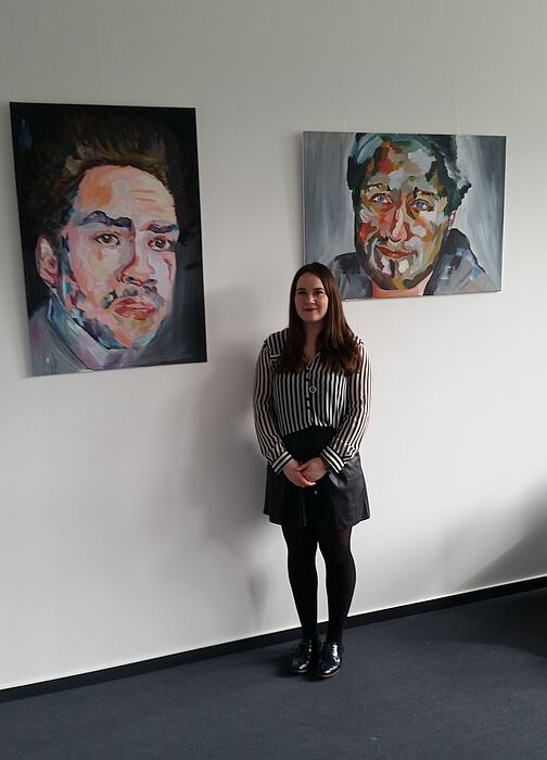 Foto: Katja Hardekopf vor einigen ihrer Porträtarbeiten auf der Ebene B2 der Uni Paderborn