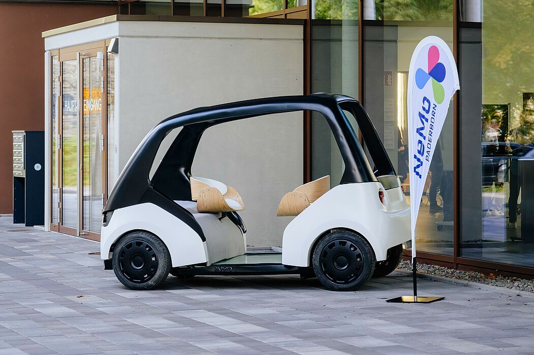 Im Vordergrund ein sogenanntes "Cab", ein Modellfahrzeug, mit dem im Projekt "Neue Mobilität Paderborn" Personen nachhaltig mobil sein sollen. Daneben eine Fahne mit dem Logo des Projekts.