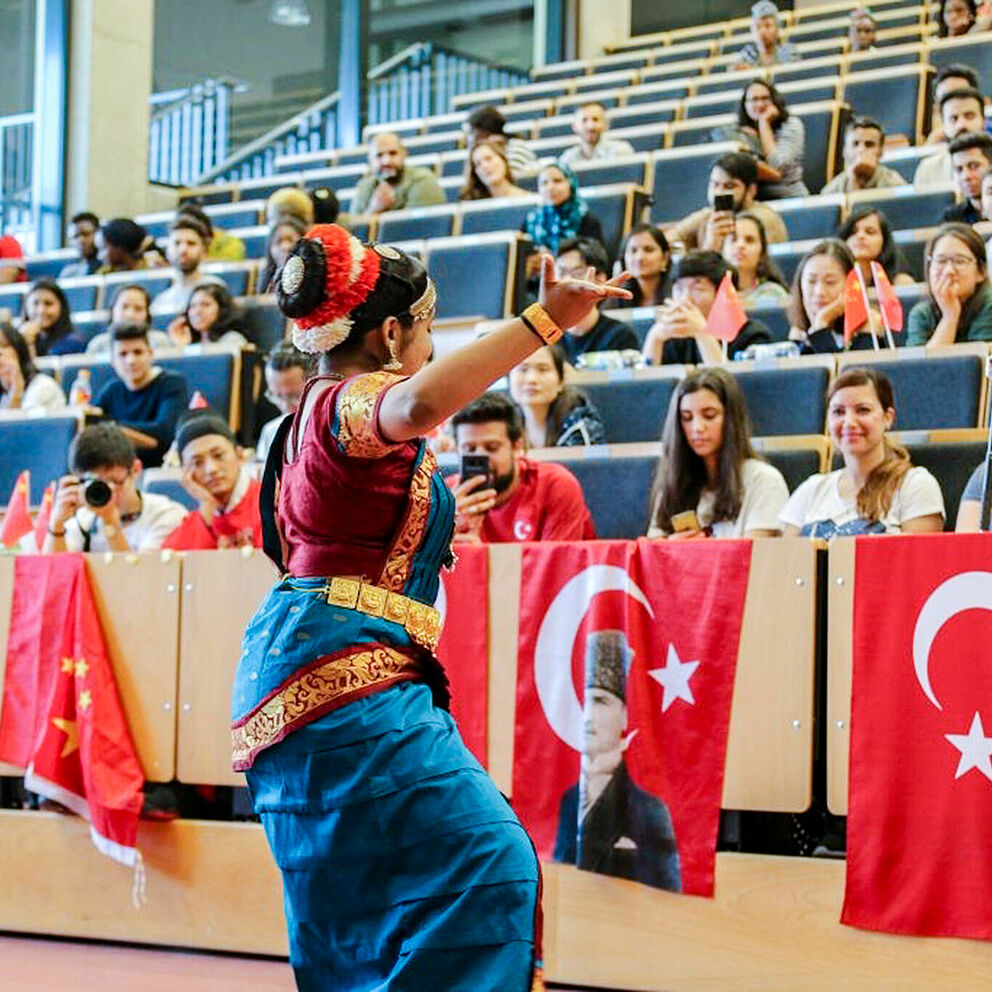 Eine Frau in traditionellem Gewand führt einen Tanz in einem Hörsaal der Universität Paderborn vor. Im Publikum sitzen junge Menschen, an den Stühlen hängen chinesische und türkische Fahnen.