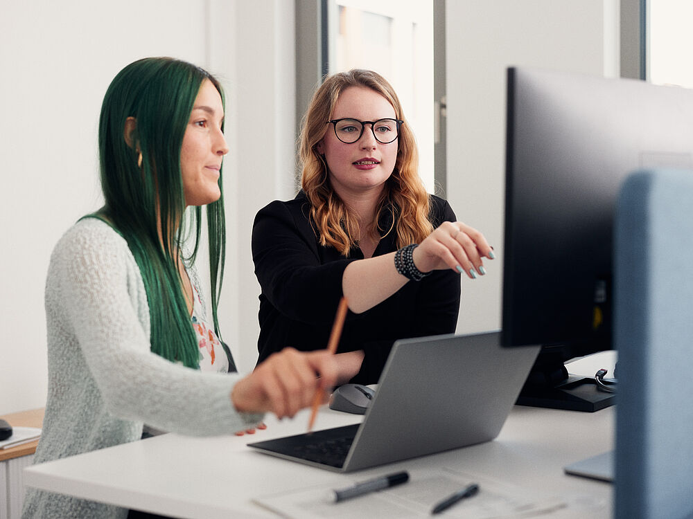 Two young female AI scientists point and look at a computer screen and discuss with each other.