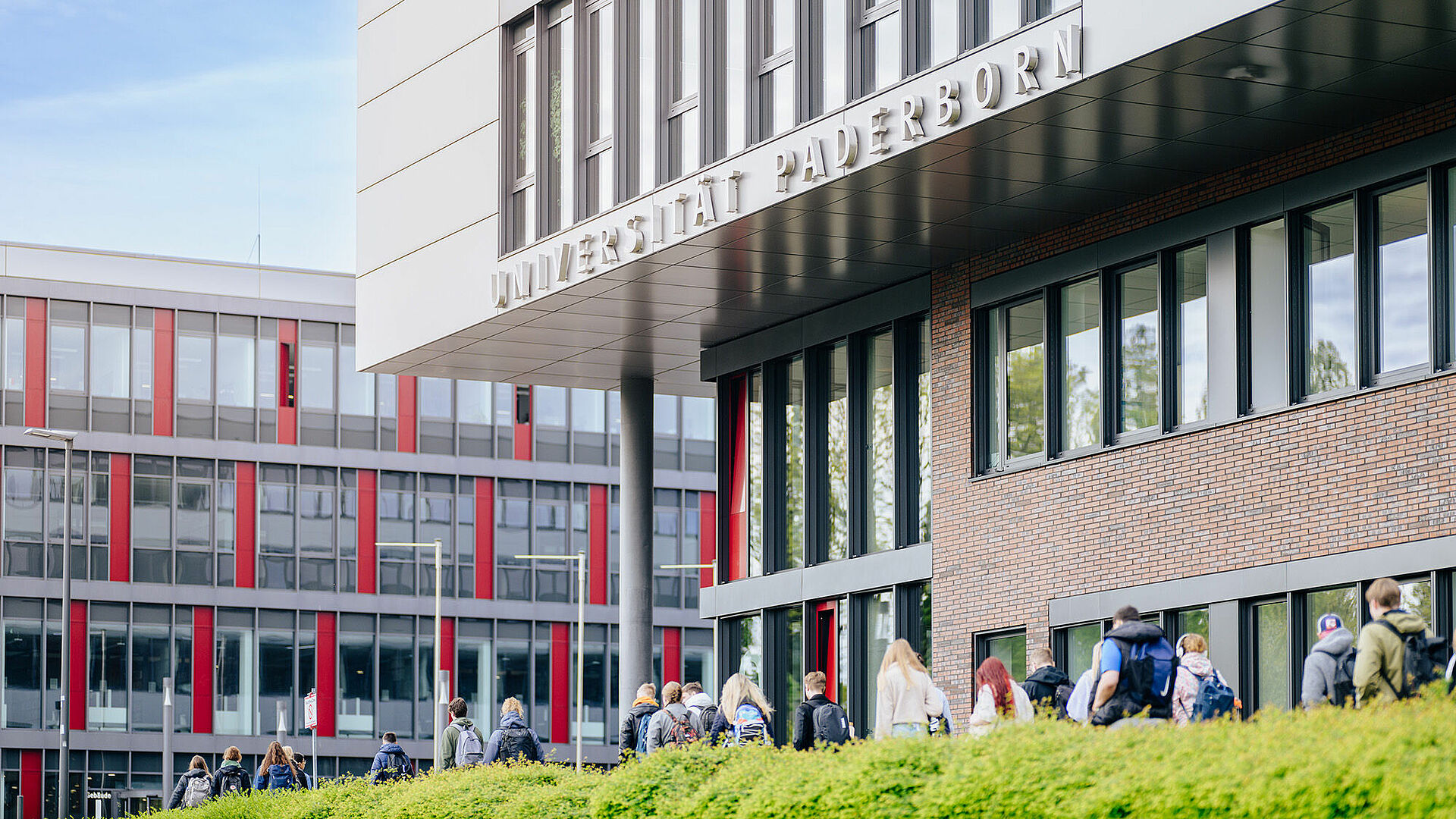 Im Vordergrund ein Teil des Gebäudes Q mit dem Schriftzug „Universität Paderborn“, vor dem mehr als 20 Studierende vorbeigehen, im Hintergrund das Gebäude I.