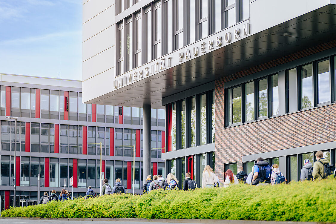 In the foreground, a part of building Q with the lettering "Universität Paderborn", in front of which more than 20 students are passing by; in the background, building I.