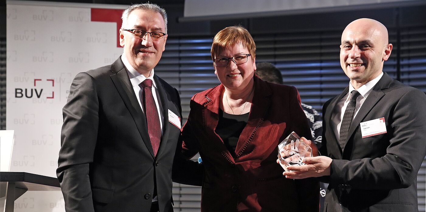 Foto (Raynet GmbH), v. li.: Der Präsident des BUV, Osman Kimil, und die Parlamentarische Staatssekretärin beim Bundesminister für Wirtschaft und Energie, Iris Gleicke, gratulieren Ragip Aydin.