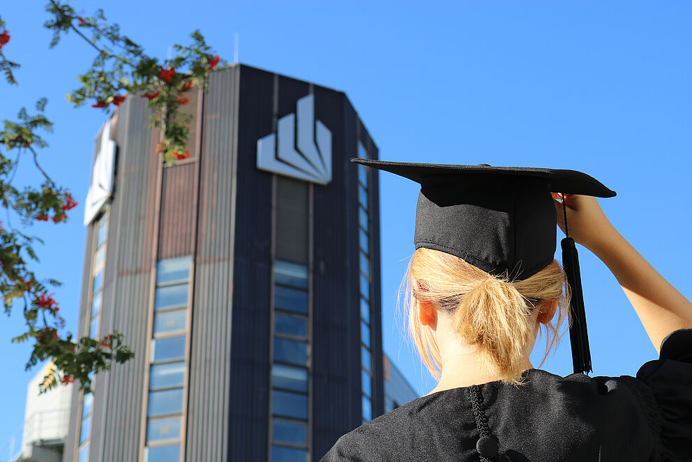 A graduate stands in front of Paderborn University wearing a gown and a doctoral hat. Her back is turned to the camera.