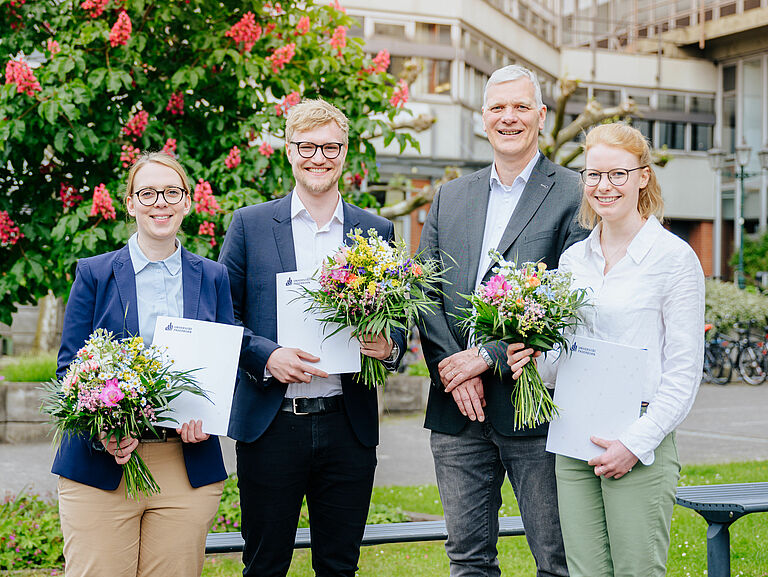 Vier Personen im Smart Casual Look stehen auf dem Campus der Universität. Die drei Preisträger:innen halten Blumensträuße und Urkunden in der Hand.