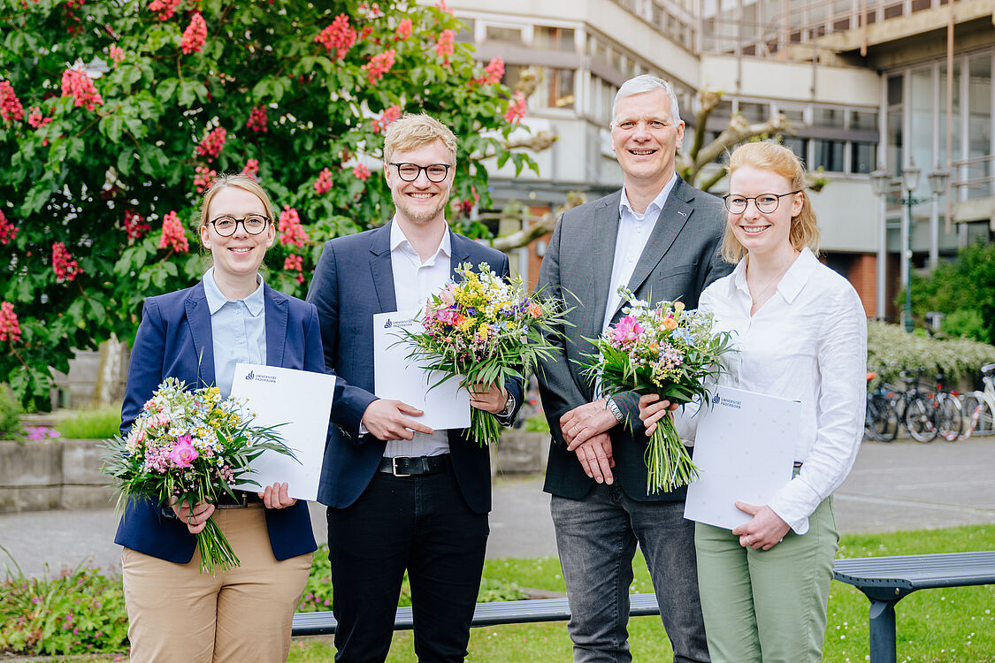 Vier Personen im Smart Casual Look stehen auf dem Campus der Universität. Die drei Preisträger:innen halten Blumensträuße und Urkunden in der Hand.