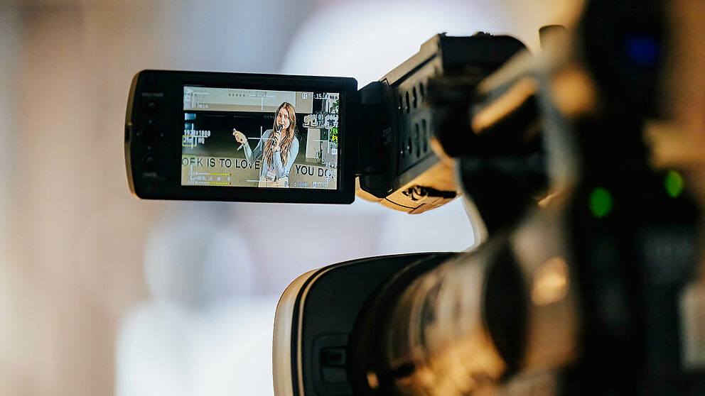 Im Vordergrund eine Kamera, auf deren Bildschirm man eine Frau erkennt, die etwas erklärt.