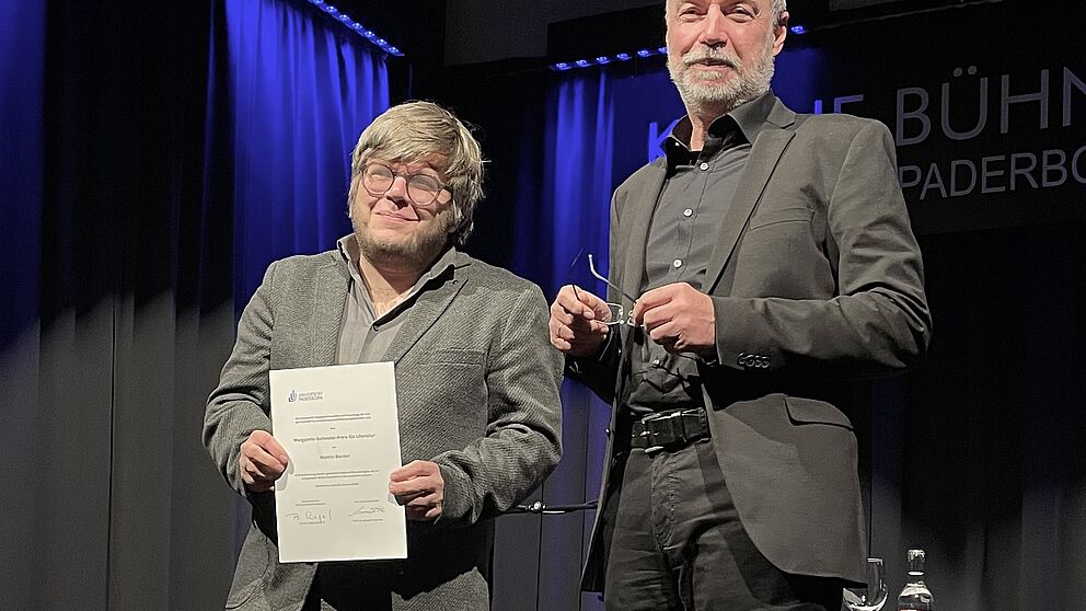 Zwei Männer stehen nebeneinander auf einer Bühne. Der links stehende Mann hält eine Urkunde. Beide Männer schauen nach unten in die Kamera.