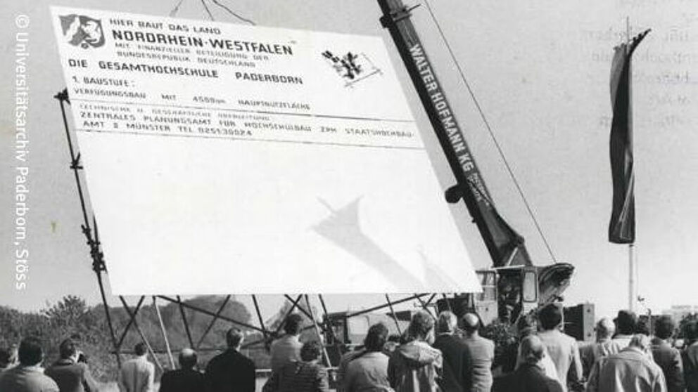 A crane erects a large sign reading "Here the state of North Rhine-Westphalia is building the Paderborn Comprehensive University". The black-and-white photograph shows several spectators from behind.
