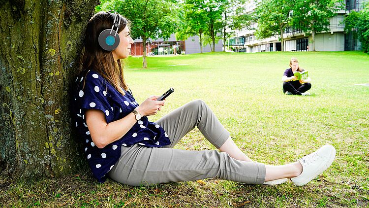 Eine Studentin sitzt auf der Wiese des Campus der Universität Paderborn, hat Kopfhörer auf und hört einen Podcast. Im Hintergrund unscharf eine Studentin, die liest.