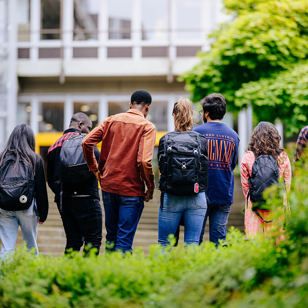 A group of students with their backs turned to the camera walks towards the entrance of Paderborn University.