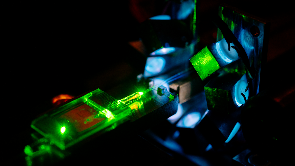 Aufnahme von einem Laser, dessen grünes Licht auf einen durchsichtigen Chip scheint. Dunkler Hintergrund