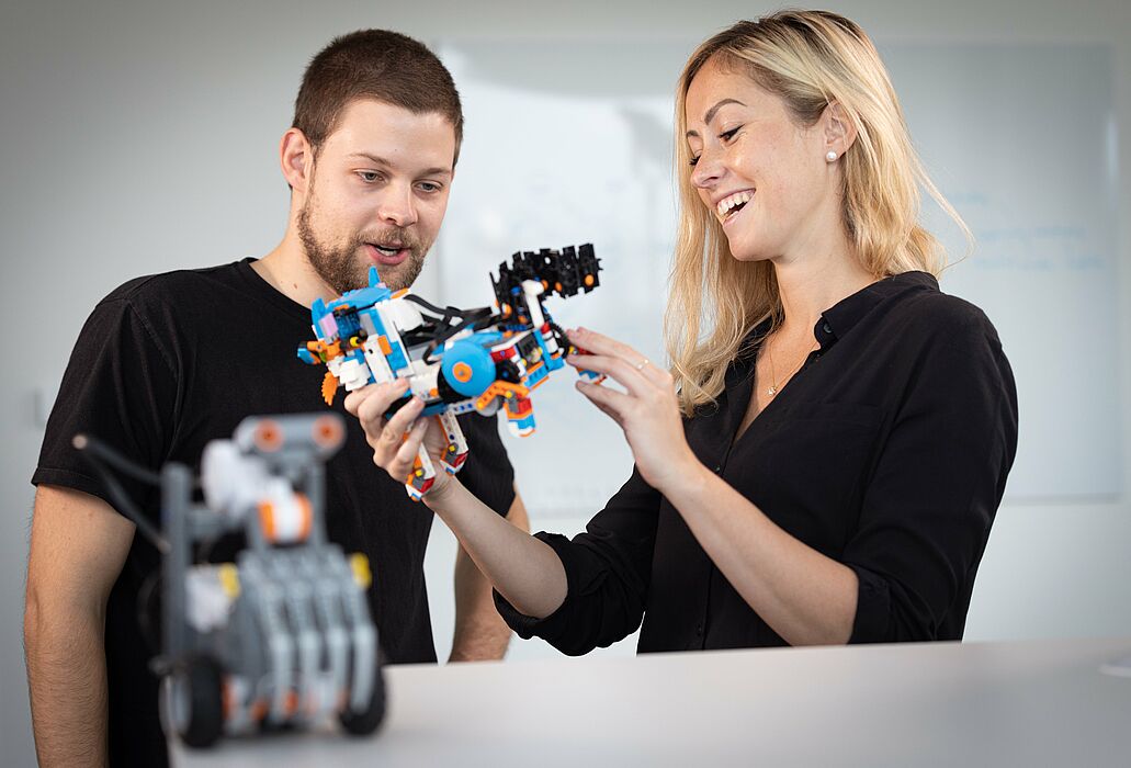 In the foreground, out of focus, a small robot, in the background a woman showing a man a robot and explaining its function.
