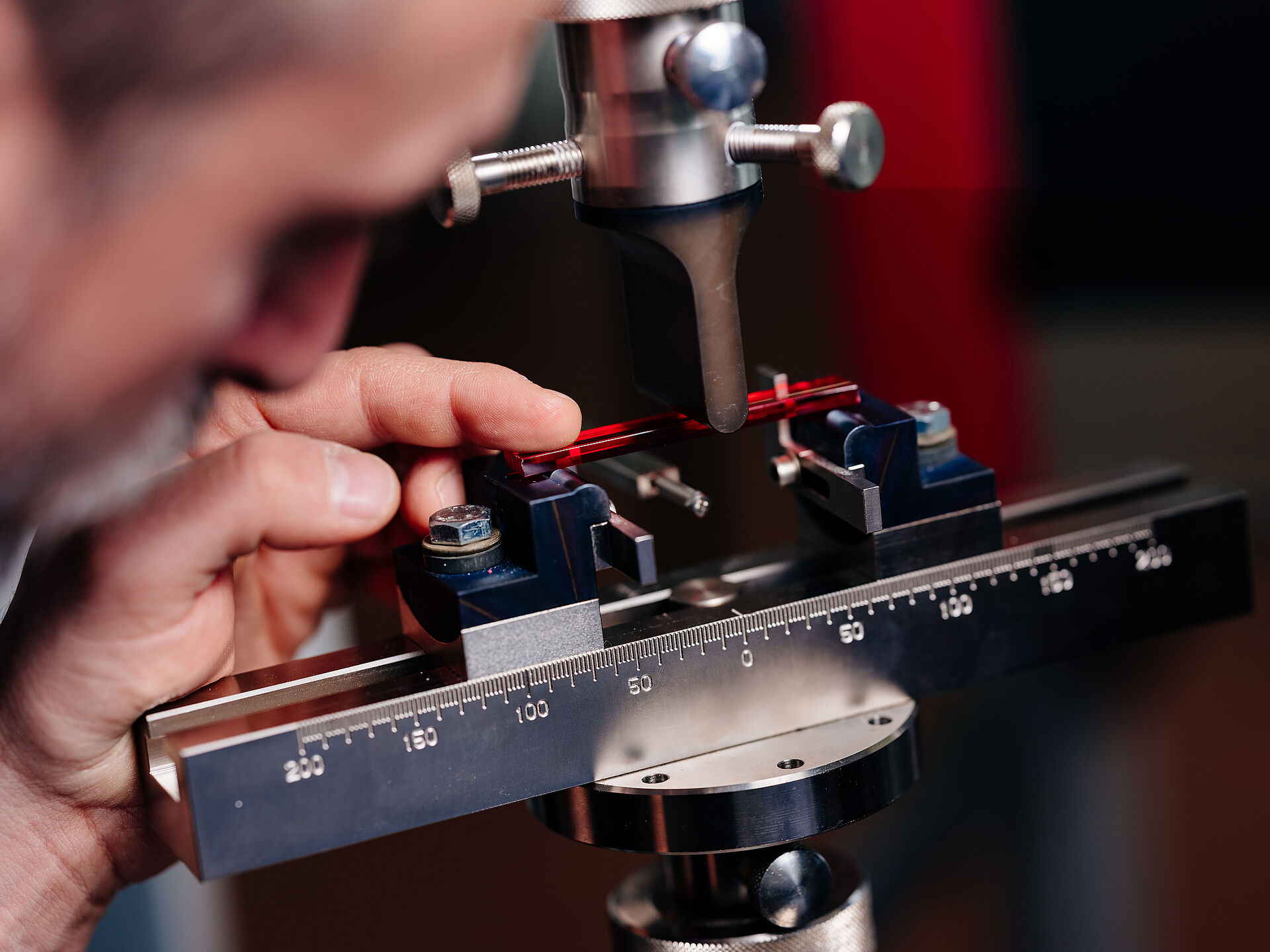 A scientist prepares to measure the deflection strength of a red colored transparent plastic strip in an apparatus