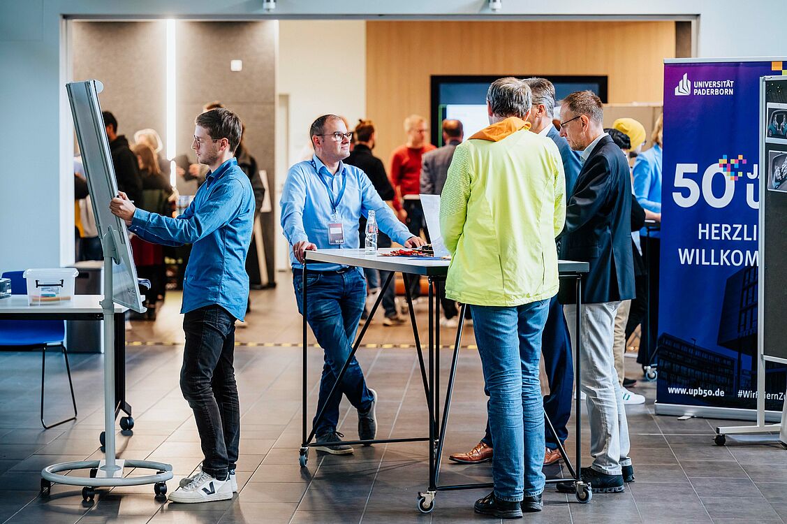 An einem Stehtisch tauscht sich ein Wissenschaftler der Universität Paderborn mit mehreren Besuchern aus. Daneben steht ein weiterer Wissenschaftler, der etwas anschaulich skizziert.