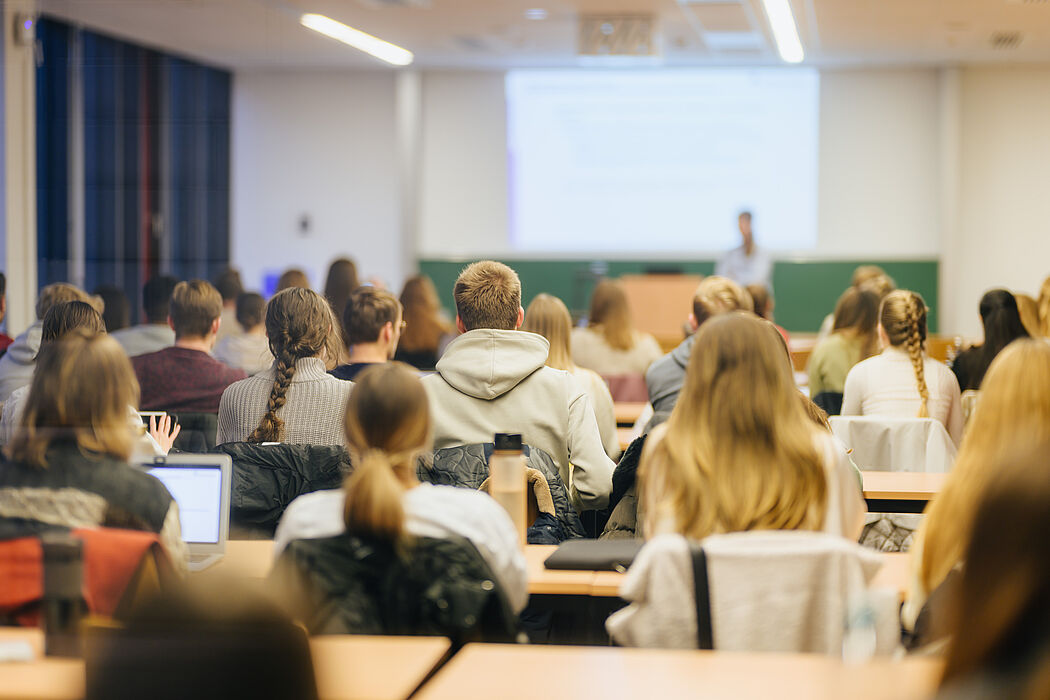 Rear view of a group of people sitting at tables in a room and looking at another person standing in front of a screen and a blackboard.