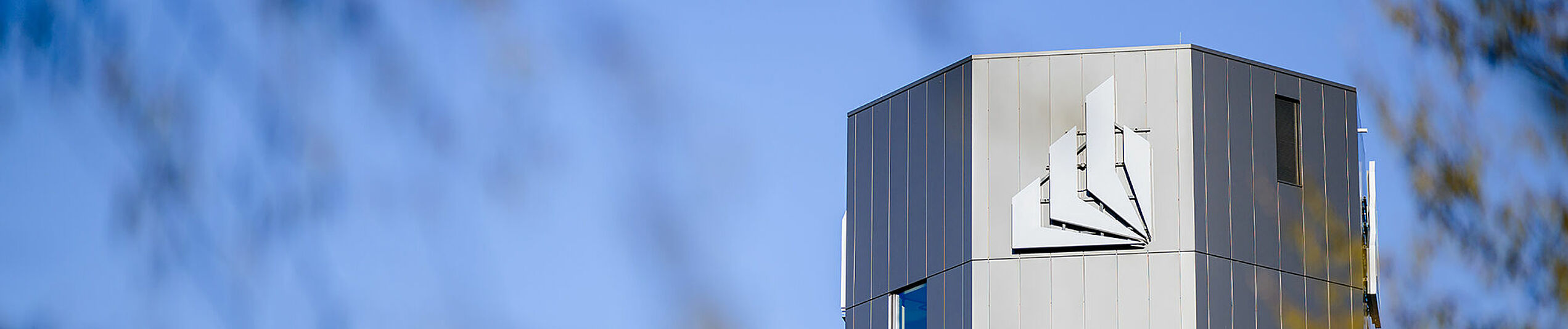 The upper part of the dark grey octahedral tower with the figurative mark of the logo of Paderborn University in front of a blue sky.
