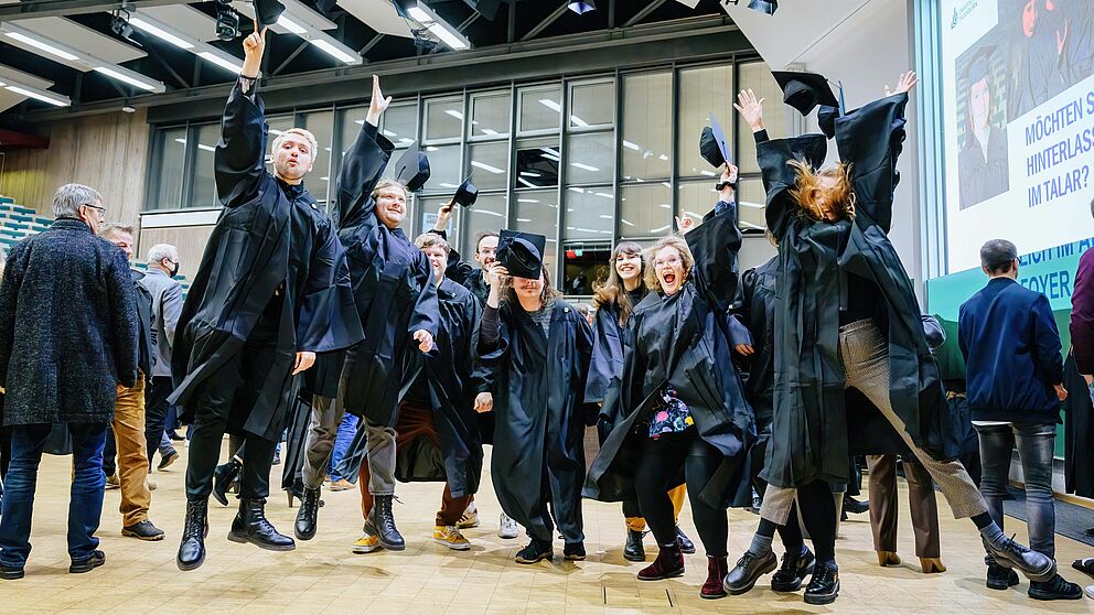 Graduates in the auditorium of Paderborn University wear gowns and throw hats into the air. 