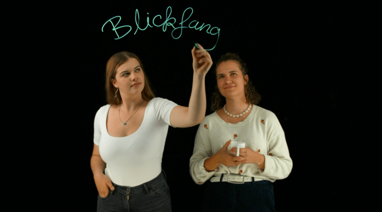 Two editors of the programme "Blickfang" write the words "Blickfang" on a pane of glass.