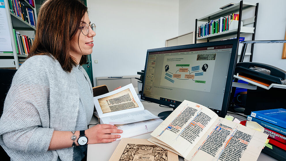 Eine Wissenschaftlerin mit einem aufgeschlagenen Buch, dass links die Abbildung eines mittelalterlichen Schriftstücks enthält, daneben zwei weitere Bücher. Dahinter ein Widescreen-Monitor mit der Anzeige einer Vortragsfolie "Maschinelle CC-Annotation"