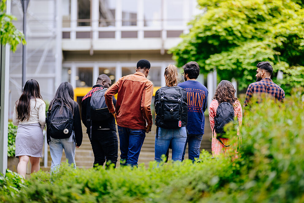 Eine Gruppe Studierender mit dem Rücken zur Kamera gedreht geht auf den Eingang der Universität Paderborn zu.