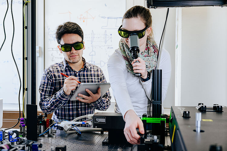 Ein junger Physiker und eine junge Physikerin stehen an einem Tisch mit technischen Geräten.