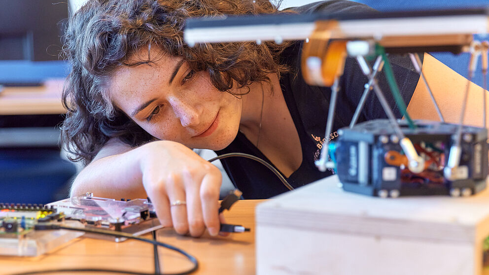 Eine junge Frau mit dunklen Haaren hält mit der Hand ein Kabel und möchte es mit einem technischen Gerät verbinden