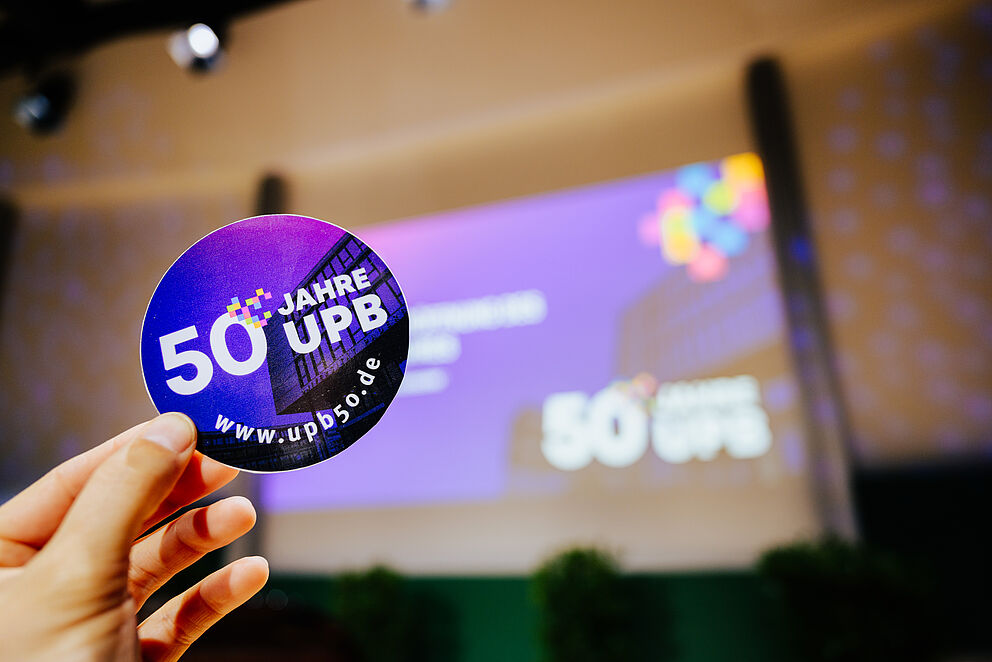 Eröffnungsfeier des 50-jährigen Jubiläums der Universität Paderborn: Eine Hand hält einen UPB50-Sticker hoch