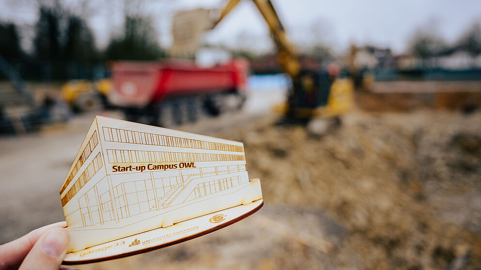 A hand holds up a wooden model of the Start-Up Campus OWL, the construction site can be seen blurred in the background.