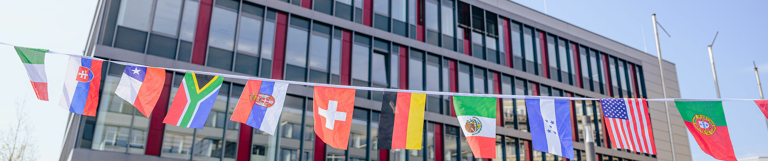 Zehn verschiedene kleine Länderflaggen aufgehängt auf einer Schnur, im Hintergrund das Gebäude I der Universität Paderborn.