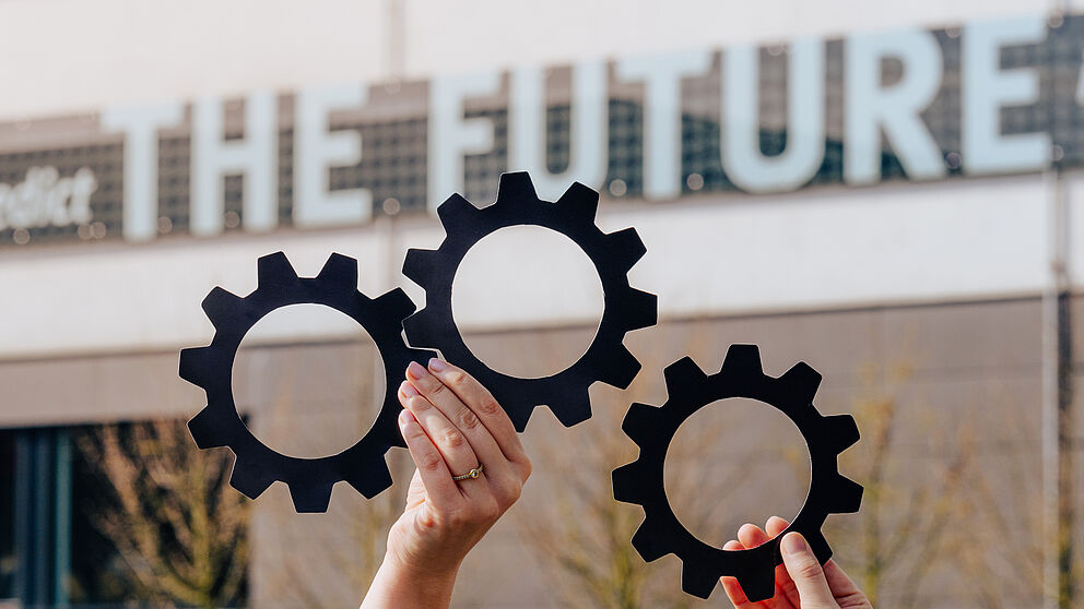 Two interlocked gears are held by one hand, a third gear engages from the right. In the background, a very blurred part of the lettering attached to Building O of Paderborn University "... best way to predict THE FUTURE" is visible in the background.