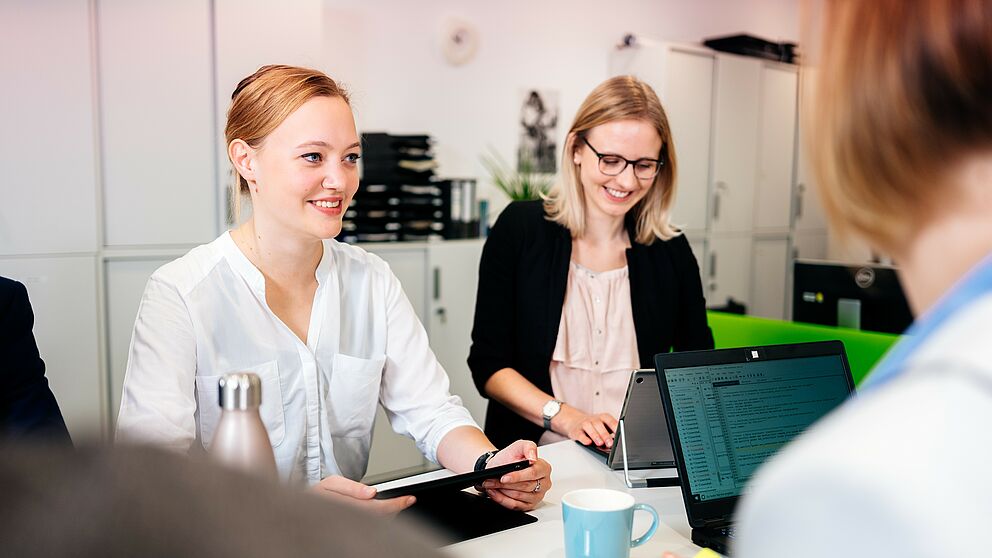 Three women sit at a workstation with laptops and tablet. 
