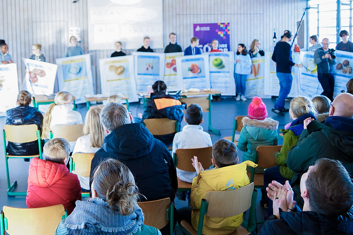 Transfer from university to school: Pupils at a performance developed by employees of Paderborn University. Adults and children watch the performance.