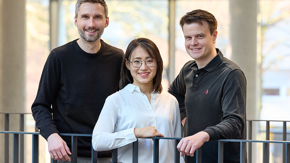 Zwei Männer und eine Frau stehen an einem Geländer und lächeln in die Kamera. 