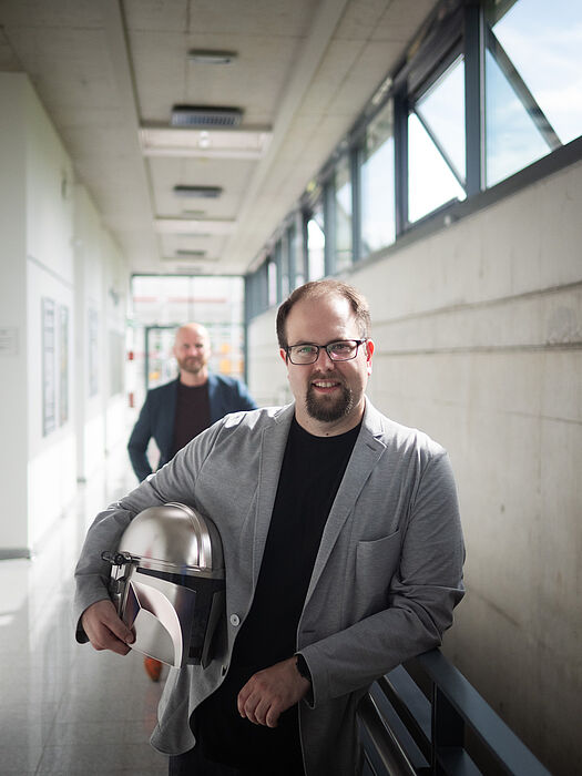 Two men are standing in a corridor, the man in front is holding a Mandalorian helmet.