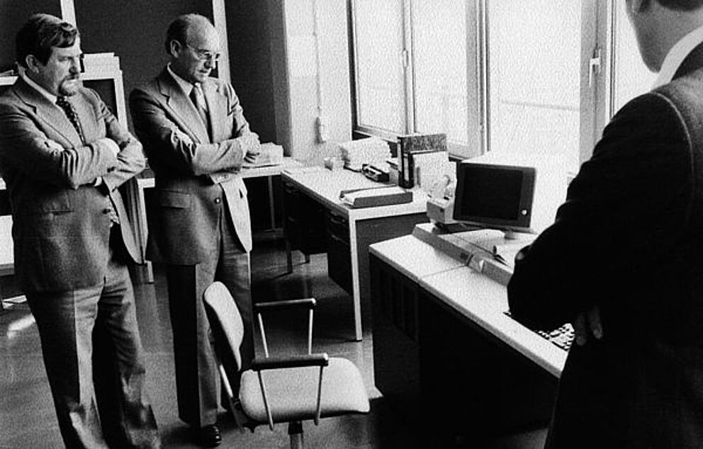 Schwarz-weiß Foto, zwei Männer stehen mit verschränkten Armen vor einem Schreibtisch und betrachten einen Computer.