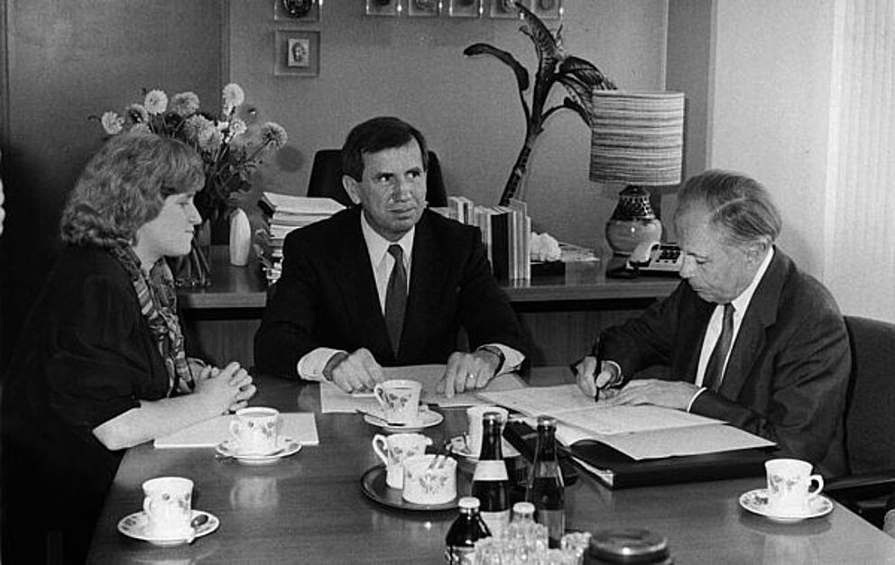 Schwarz-weiß Foto, drei Personen sitzen an einem Tisch, vor ihnen liegen Dokumente, der Mann rechts unterschreibt seine Dokumente gerade.