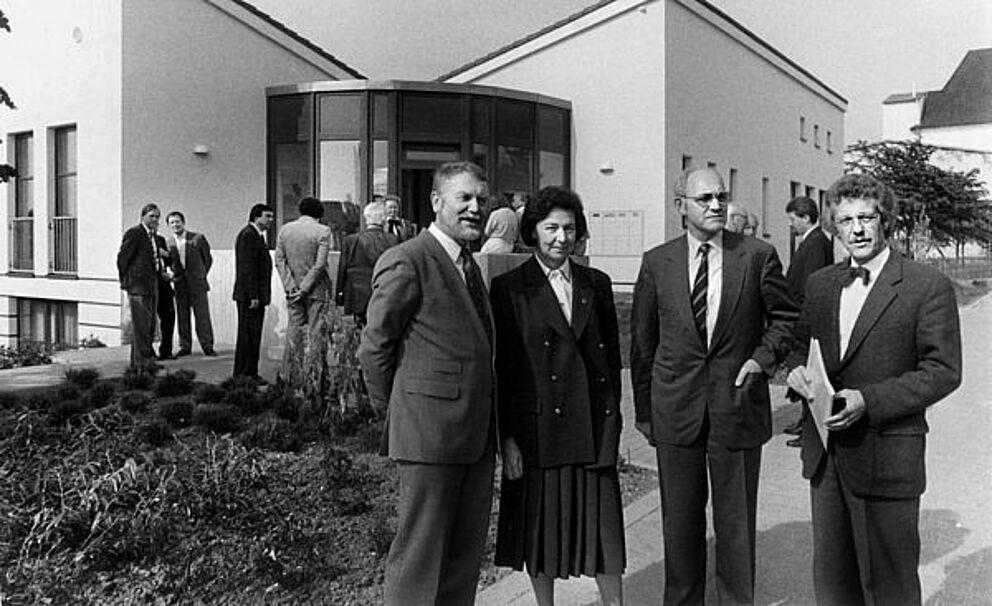 Schwarz-weiß Foto, vier Personen stehen vor einem Gebäude, im Hintergrund sind mehrere Menschen versammelt.