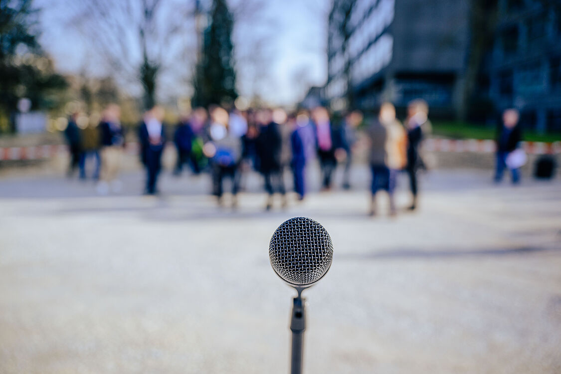 Im Vordergrund steht ein Mikrofon, im Hintergrund unscharf mehrere Leute auf dem Campus der Universität Paderborn, die zuhören.