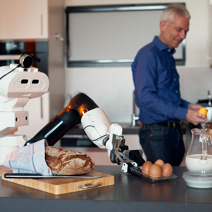 Ein Roboter bereitet Frühstück zu. Im Hintergrund steht ein Mensch.