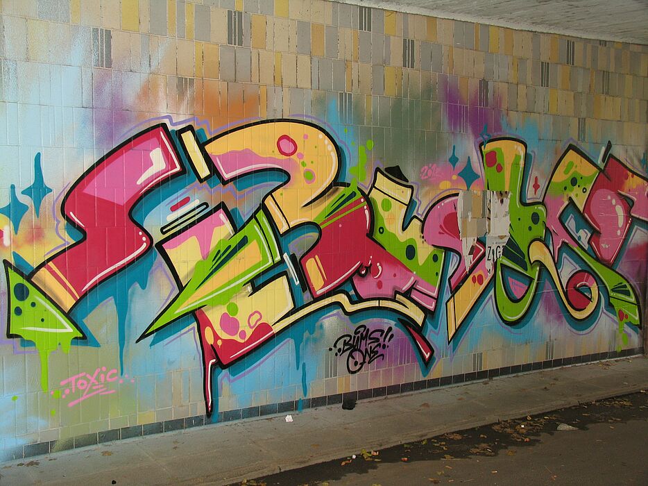 Abbildung (Universität Paderborn): Writings bzw. Styles sind aufwändiger gesprühte, mehrfarbige bildhafte Graffiti, die im Vergleich zum Throw-Up oft weitere Stilmittel wie einen farbigen Hintergrund (Background), Binnenkonturen (Inlines), Bewegungslin