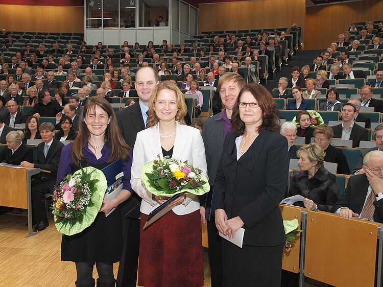 Die vier Lehrpreisträger:innen stehen neben der Vizepräsidentin vorne im Hörsaal und halten Blumensträuße in der Hand. 