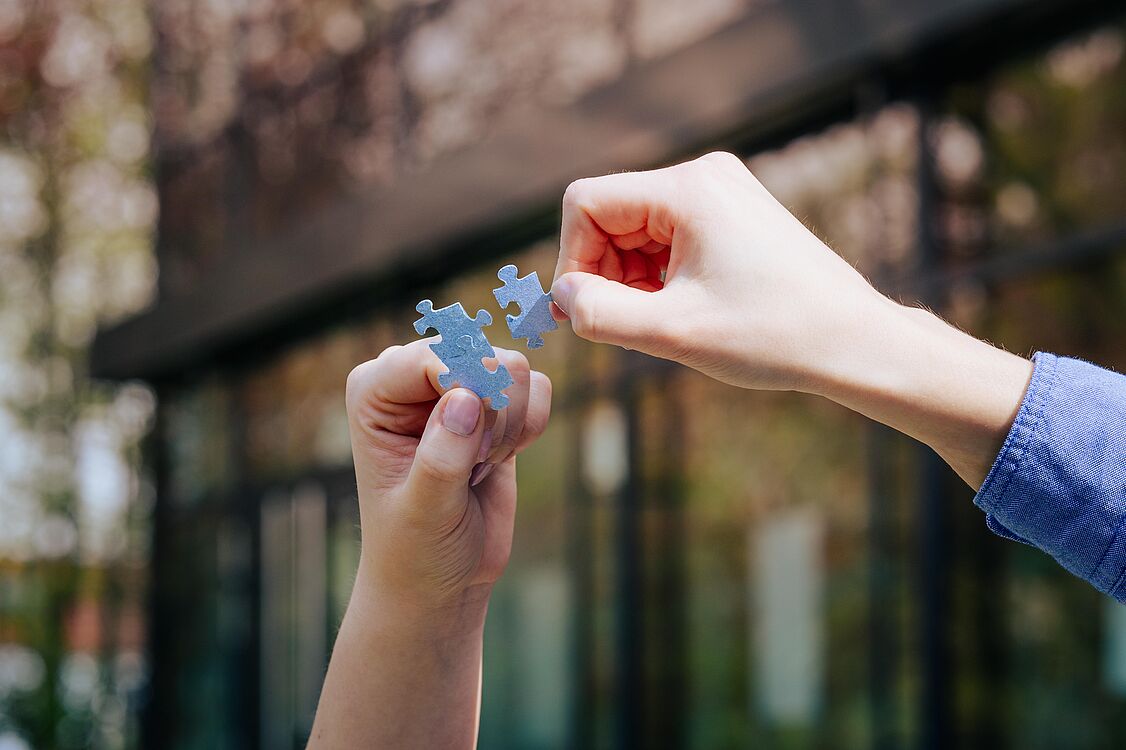 A left hand holds two interlocking gray puzzle pieces, a right hand holds a single gray puzzle piece. In the background, out of focus, the exterior view of the refectory of Paderborn University.