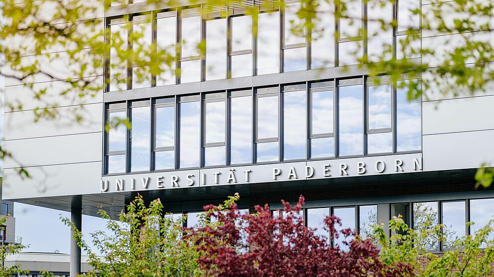 Im Hintergrund scharf das Gebäude Q der Universität Paderborn, im Vordergrund unscharf Bäume.