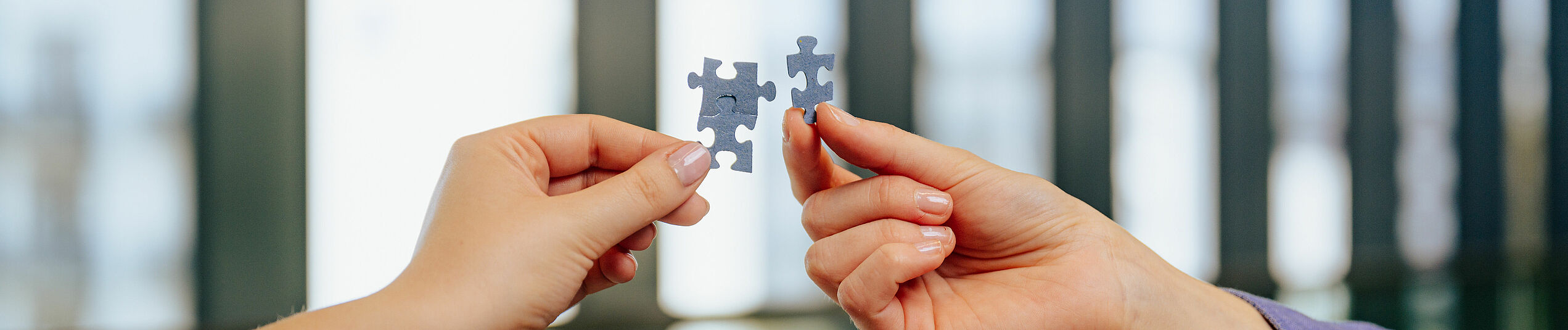 Eine linke Hand hält zwei ineinander verhakte graue Puzzle-Teile, eine rechte Hand ein einzelnes graues Puzzle-Teil