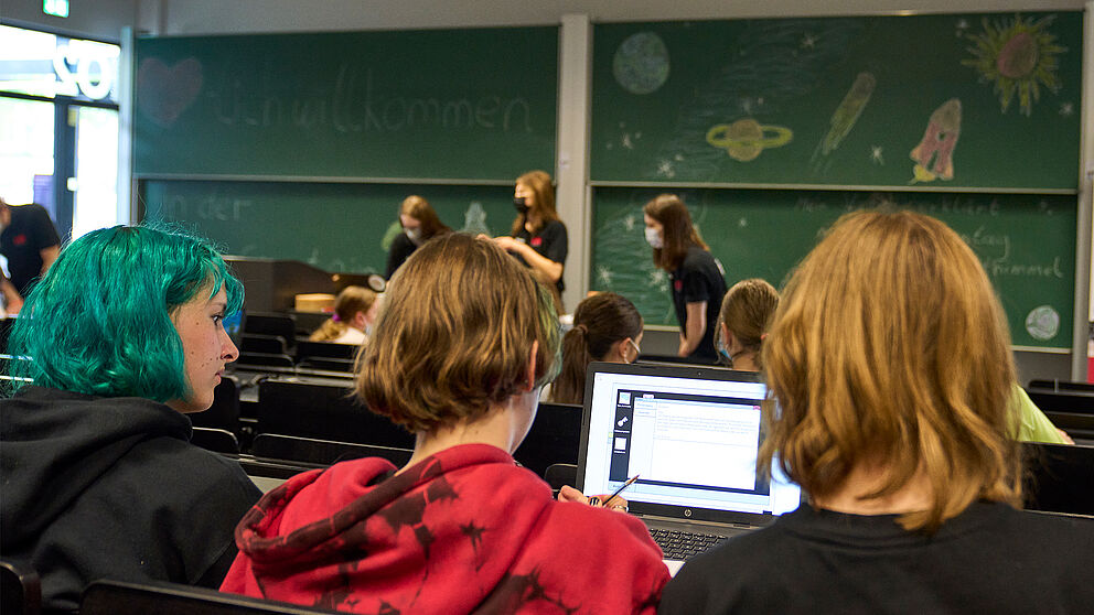 Drei Schülerinnen sitzen in Hörsaal O2 gemeinsam an einem Laptop. Im Hintergrund sind weitere Teilnehmer der Vorlesung zu sehen. 