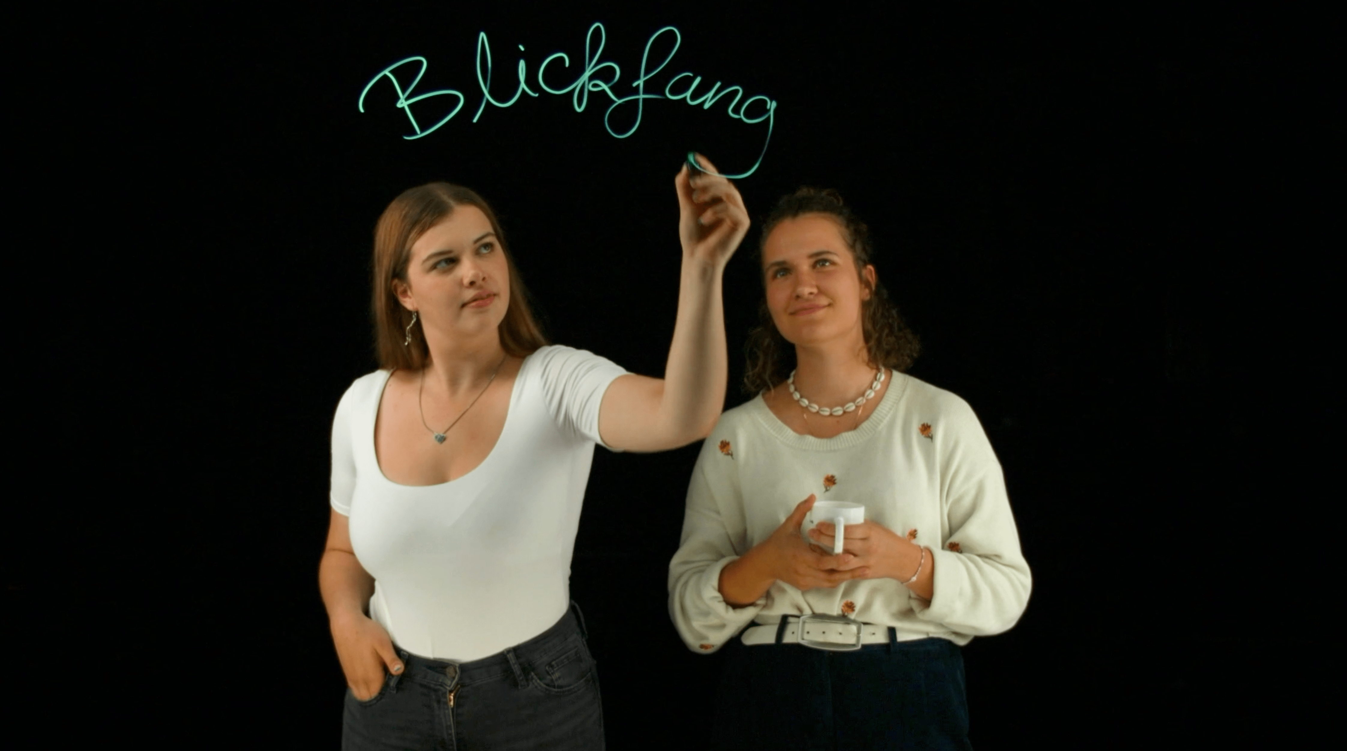 Zwei Redakteurinnen der Sendung „Blickfang“ schreiben auf eine Glasscheibe den Schriftzug „Blickfang“.